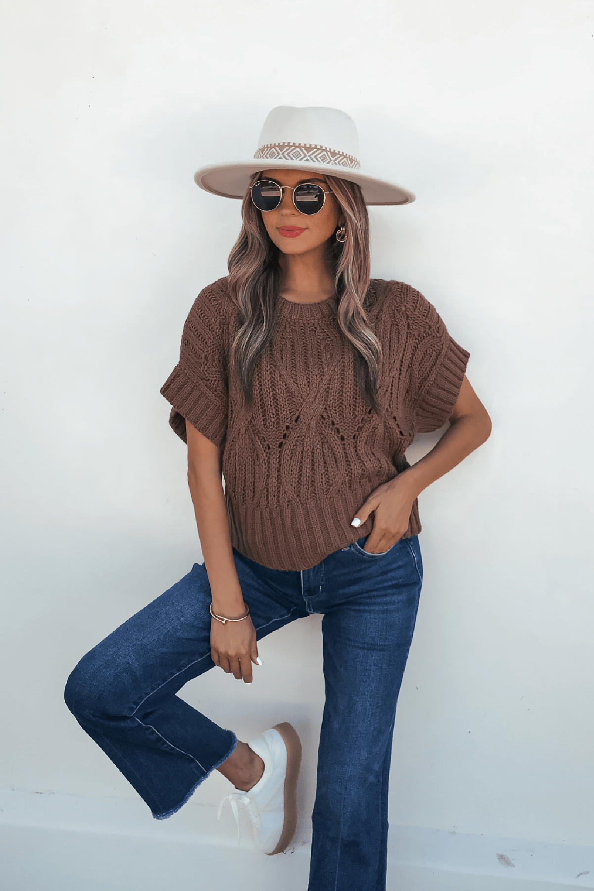 Woman in a dark brown crochet knit sweater, blue jeans, white sneakers, sunglasses, and beige hat poses against a white wall.