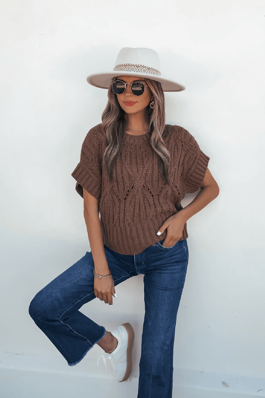 Woman in a dark brown crochet knit sweater, blue jeans, white sneakers, sunglasses, and beige hat poses against a white wall.