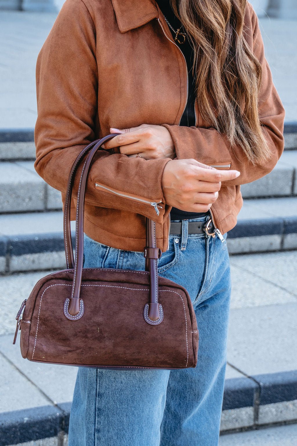 Woman in a brown suede jacket and jeans holds the Dark Brown Faux Suede Baguette Bag while standing on outdoor steps.