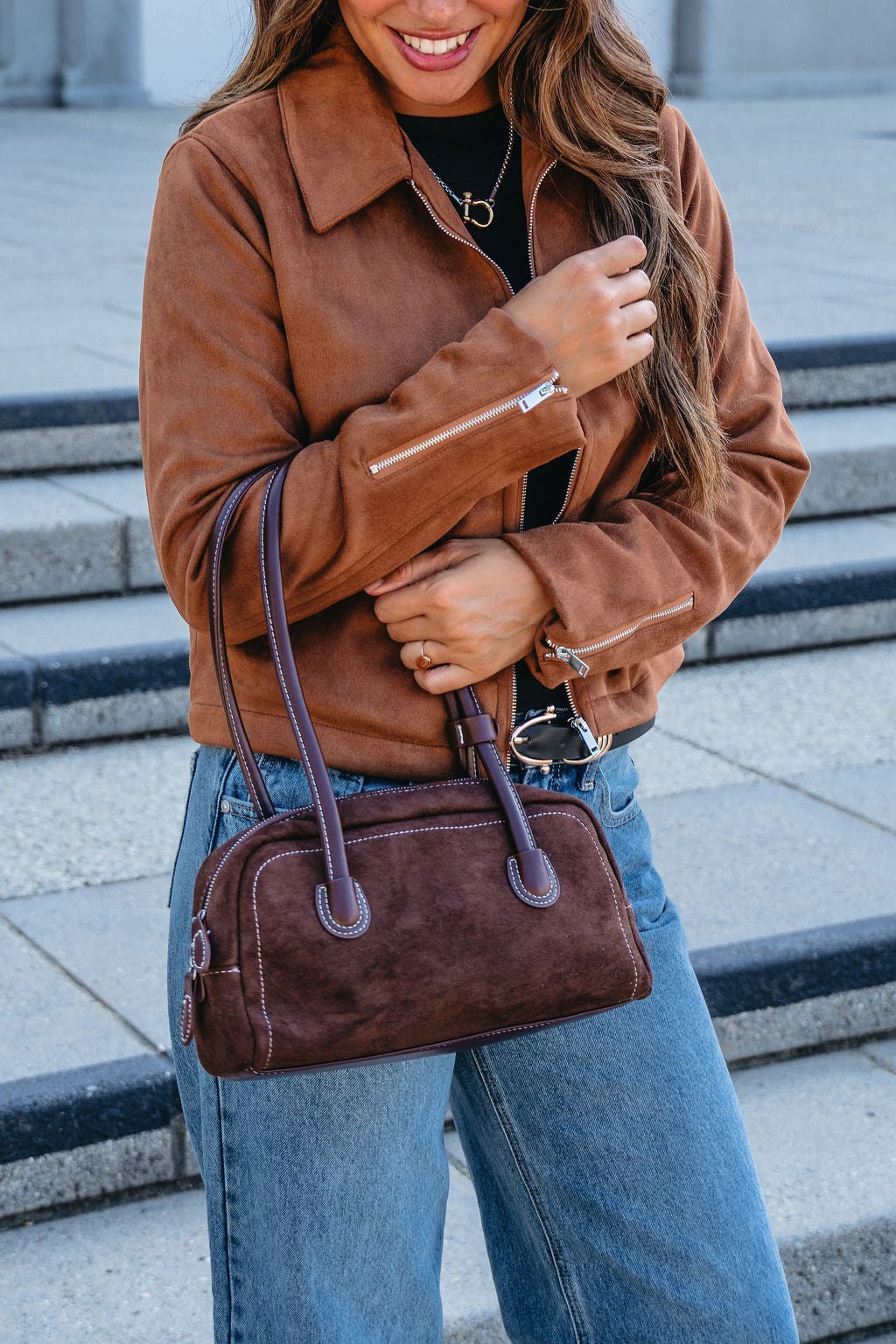 A woman stands on outdoor steps in jeans and a brown suede jacket, holding the Dark Brown Faux Suede Baguette Bag.