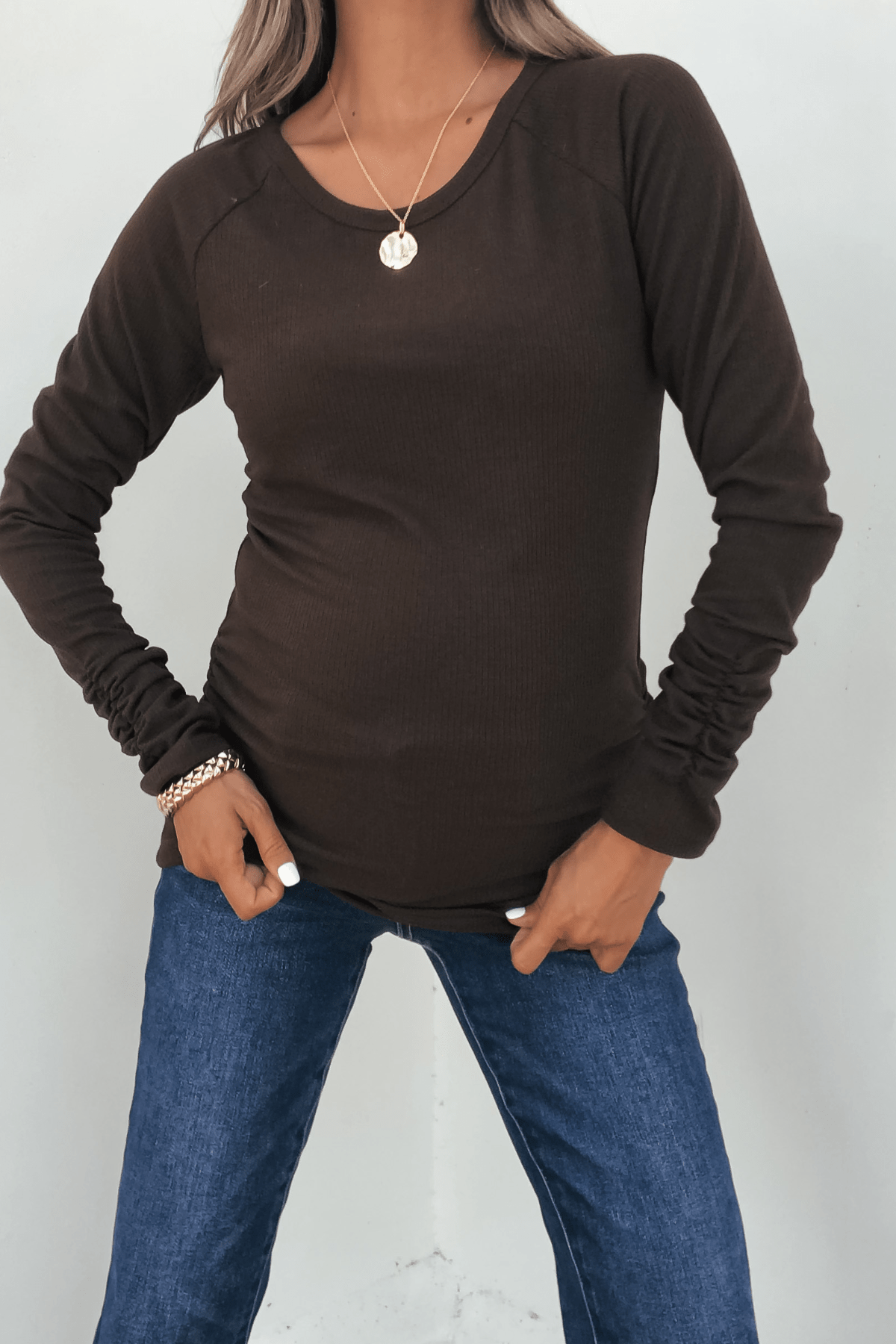Person wears a Dark Brown Ruched Sleeve Ribbed Top, blue jeans, gold necklace, and bracelets with hands in jean pockets.