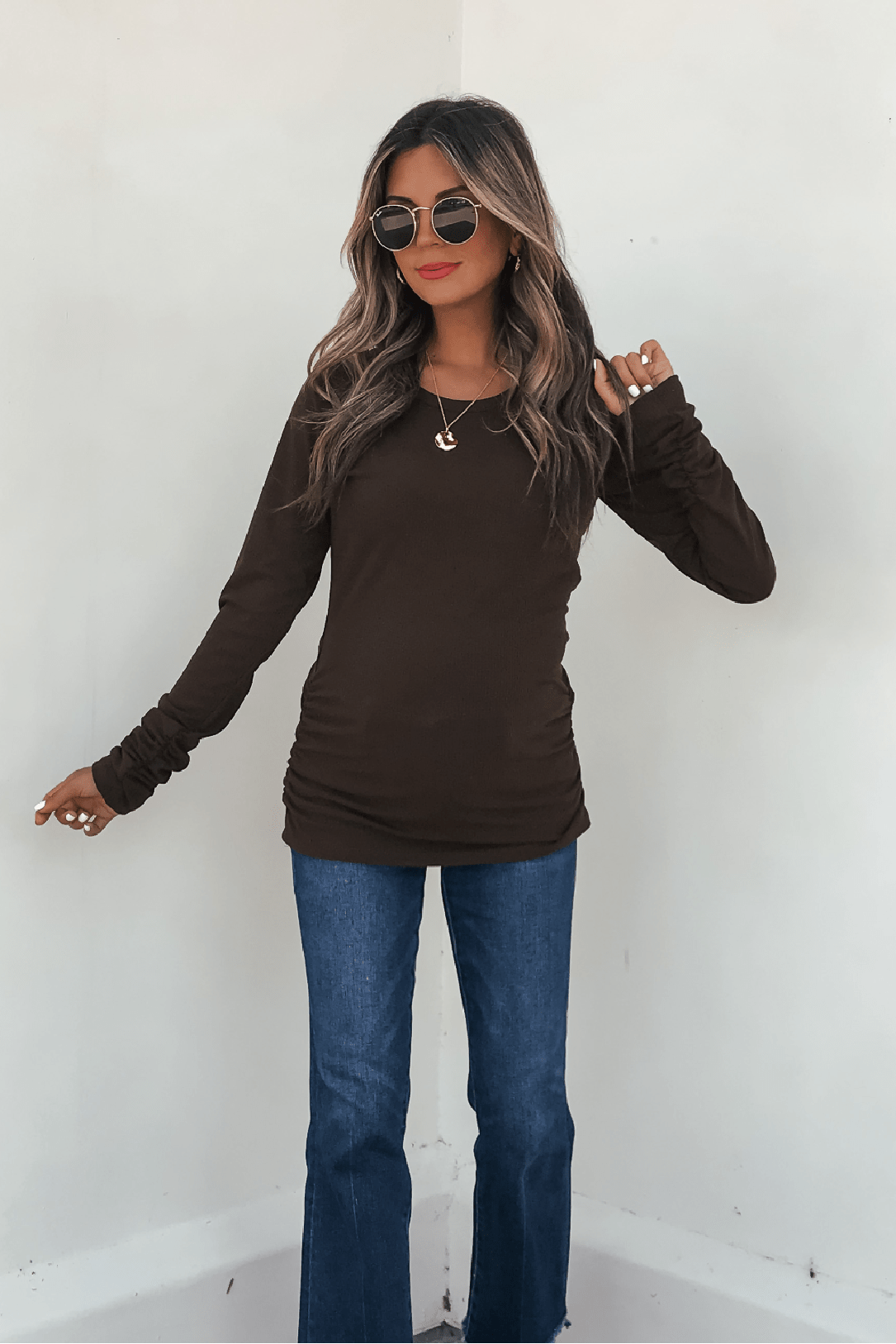 Woman in sunglasses, blue jeans, gold jewelry, and a Dark Brown Ruched Sleeve Ribbed Top stands by a white wall.