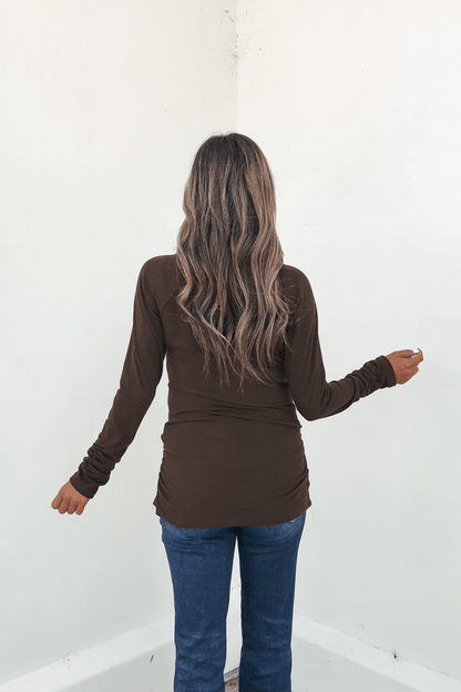 A woman with long wavy hair wears a Dark Brown Ruched Sleeve Ribbed Top and blue jeans, facing a white wall with arms slightly out.