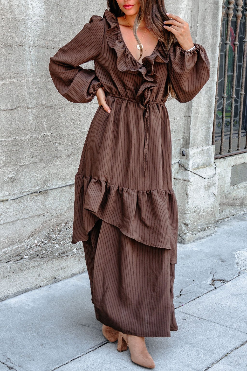 Woman in a dark brown striped ruffle tiered maxi dress stands by a textured stone wall.