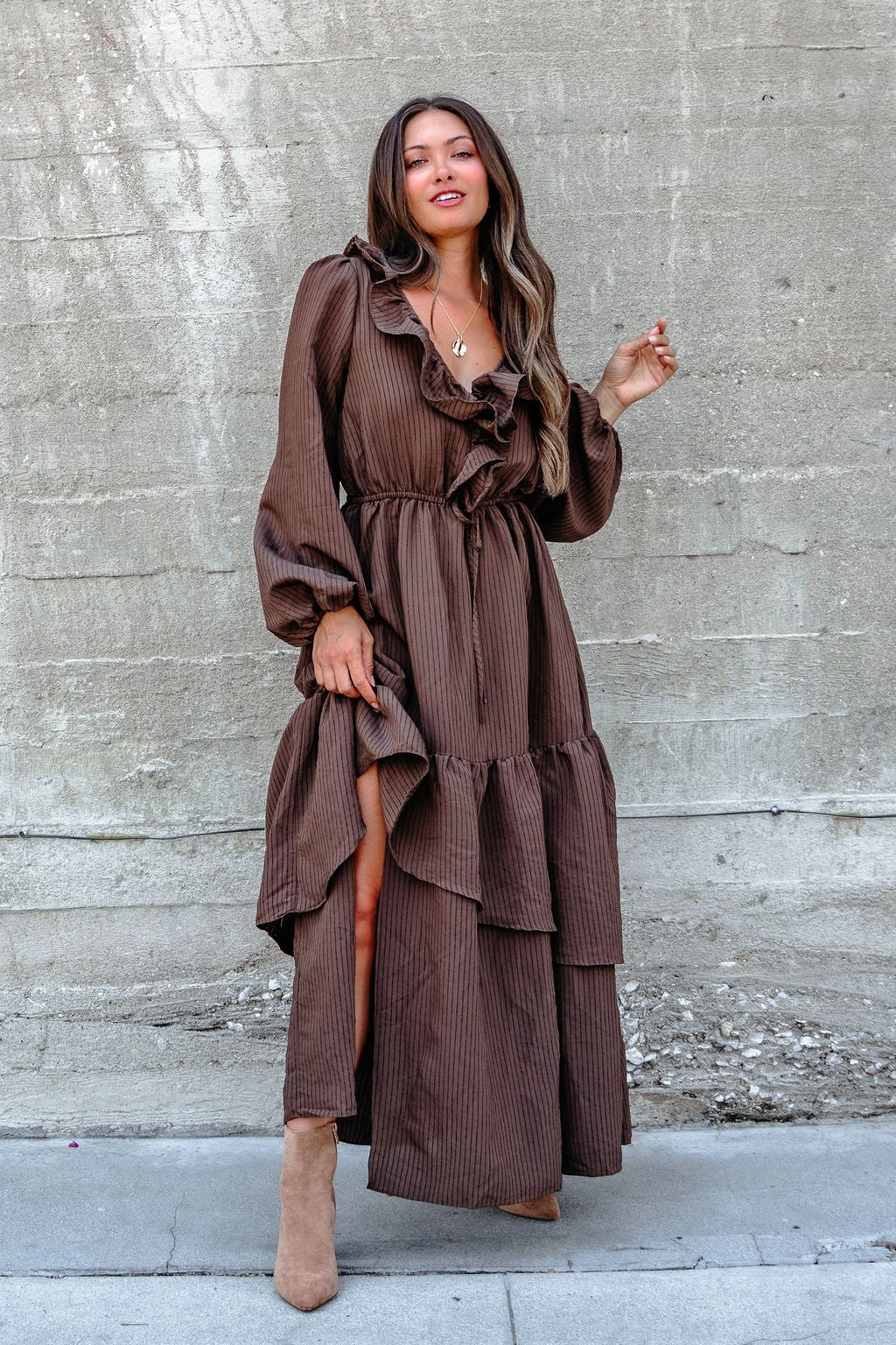 A woman in a Dark Brown Striped Ruffle Tiered Maxi Dress stands by a textured concrete wall.