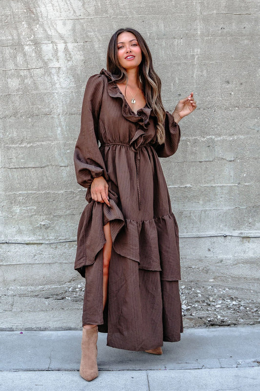 A woman in a Dark Brown Striped Ruffle Tiered Maxi Dress stands by a textured concrete wall.