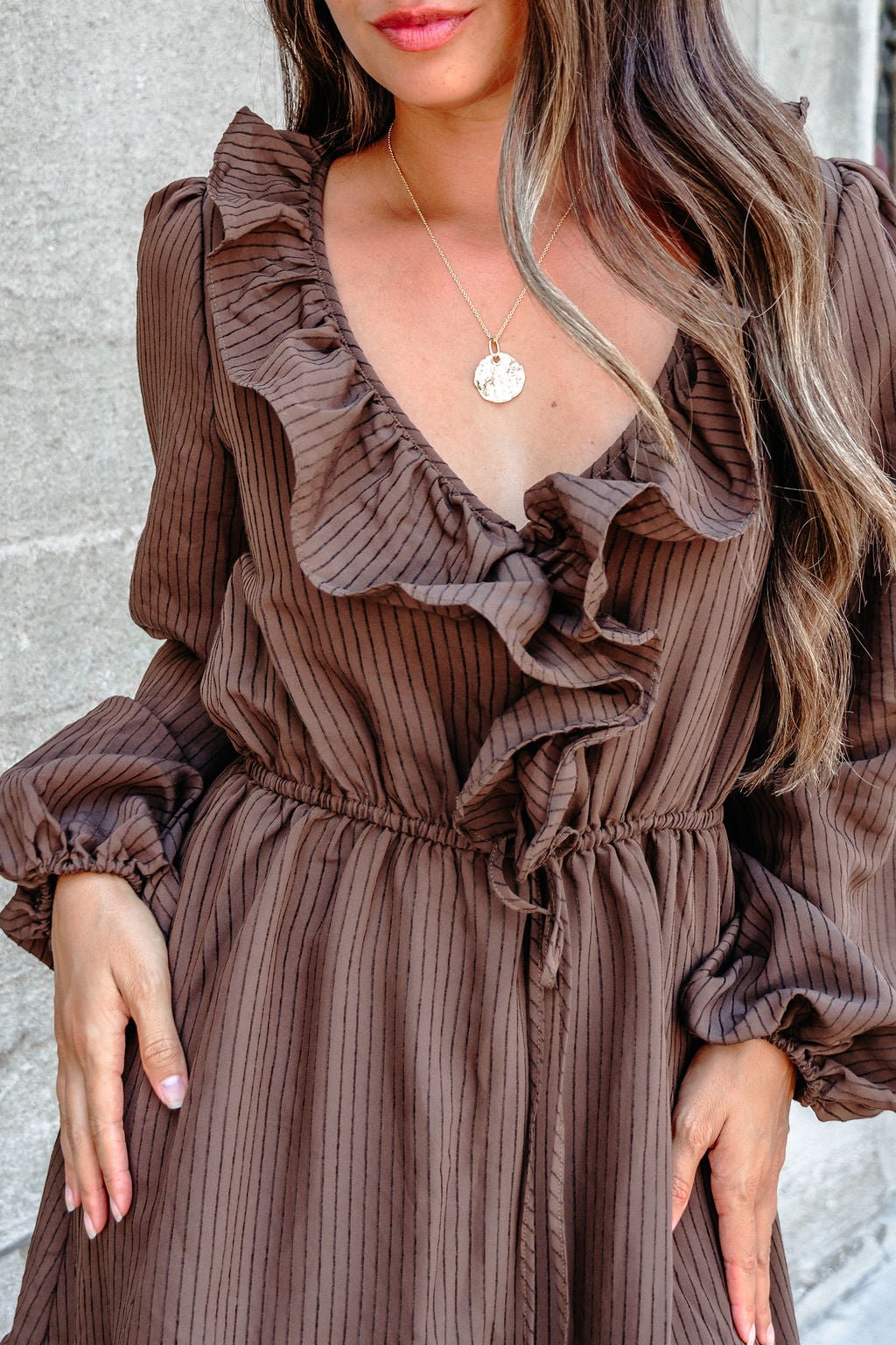 Woman in a Dark Brown Striped Ruffle Tiered Maxi Dress with long sleeves and gold necklace, standing by a gray wall.