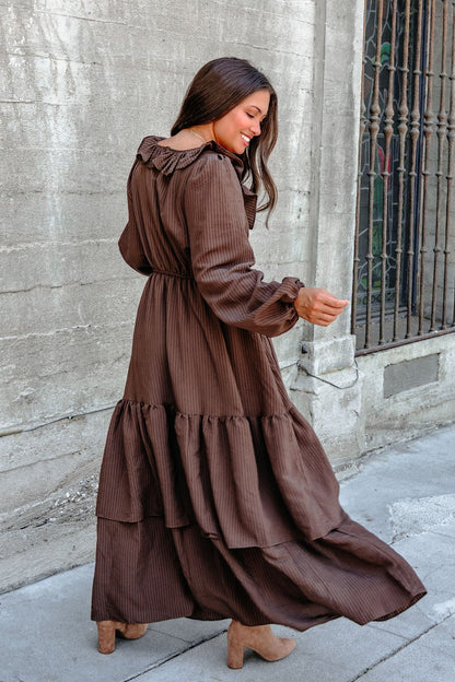 Woman in a Dark Brown Striped Ruffle Tiered Maxi Dress and tan boots, smiling as she turns beside a textured concrete wall.
