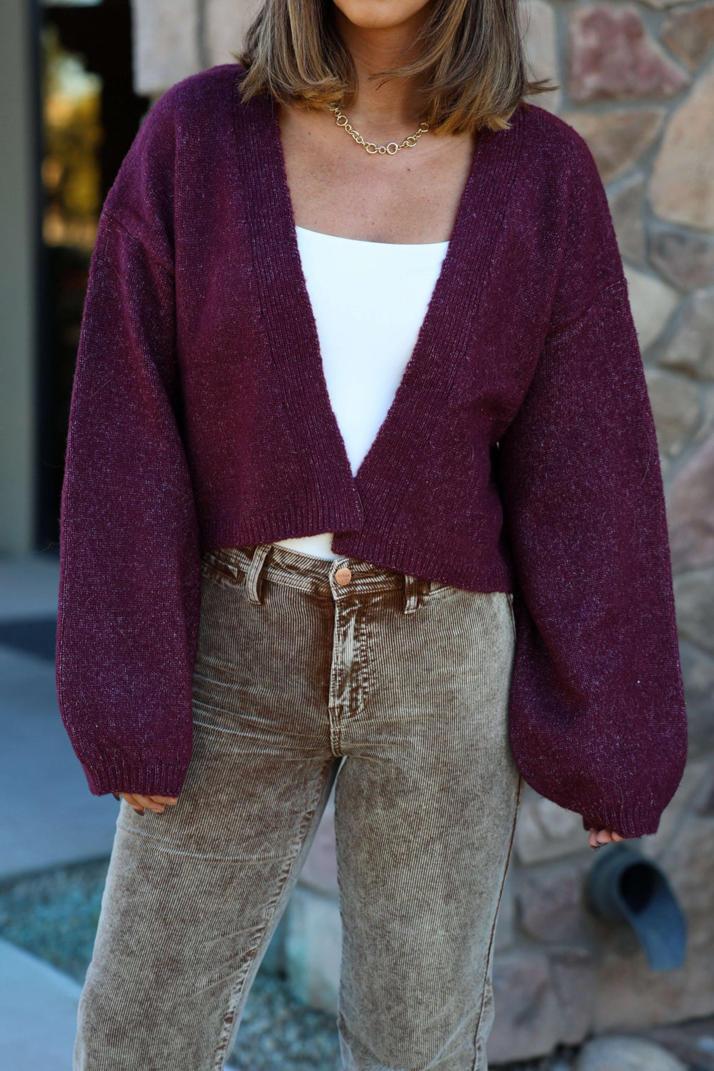 Woman stands outdoors by a stone wall wearing the Dark Cherry Open Front Cardigan - FINAL SALE over a white top and tan pants.
