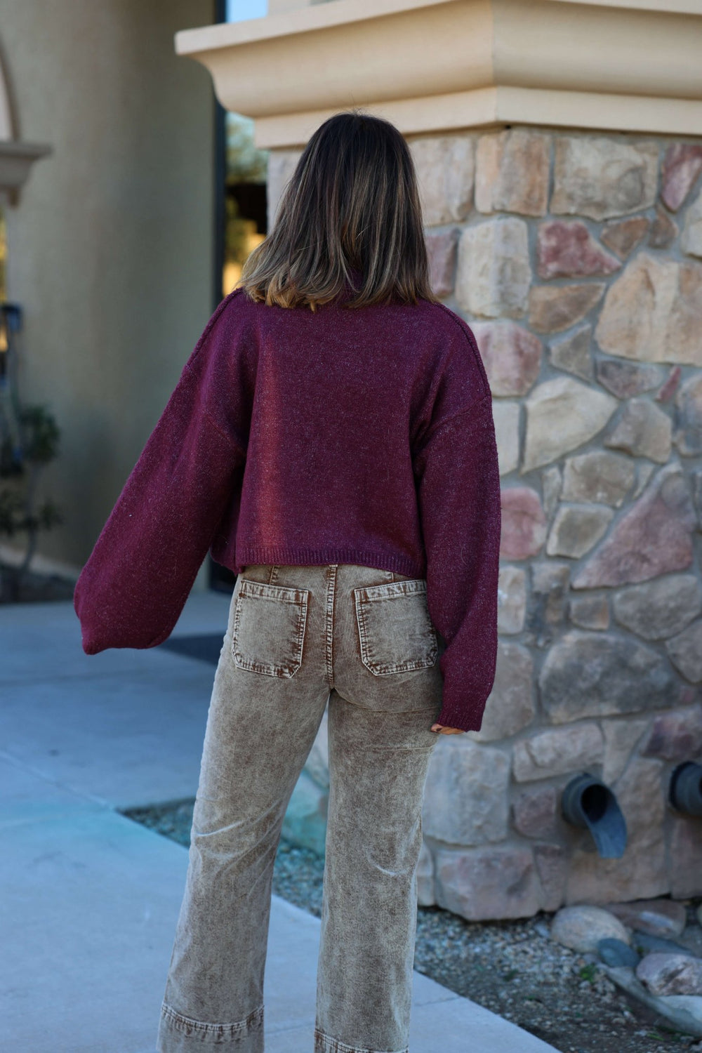 A person wears the Dark Cherry Open Front Cardigan - FINAL SALE and beige pants, seen from behind outside by a stone wall.