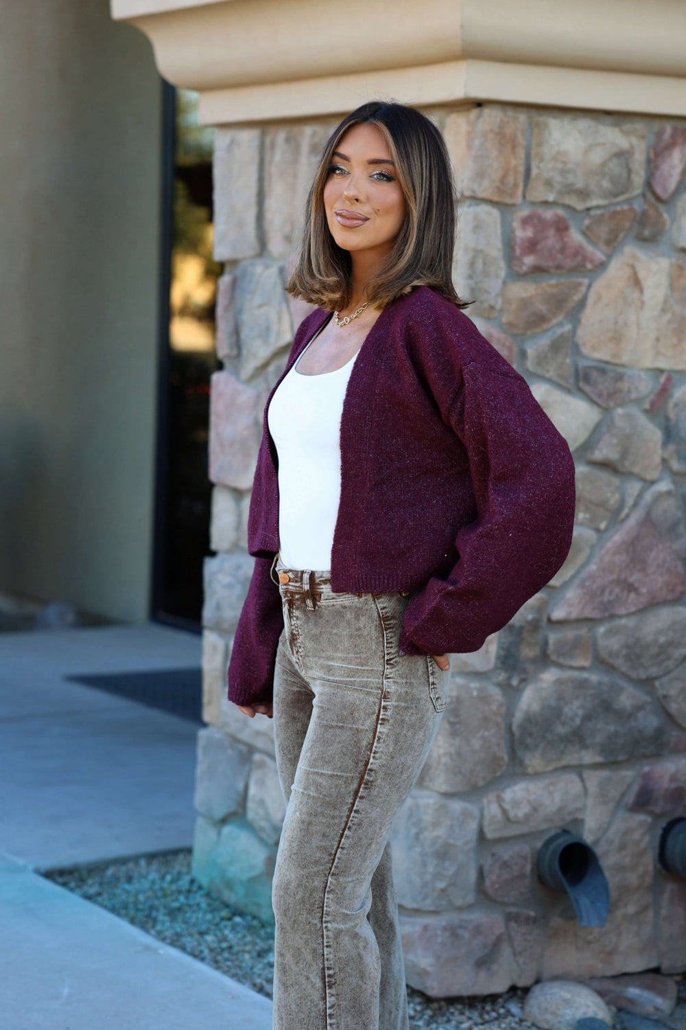 Smiling woman in a Dark Cherry Open Front Cardigan - FINAL SALE, white top, and beige jeans stands by a stone wall.