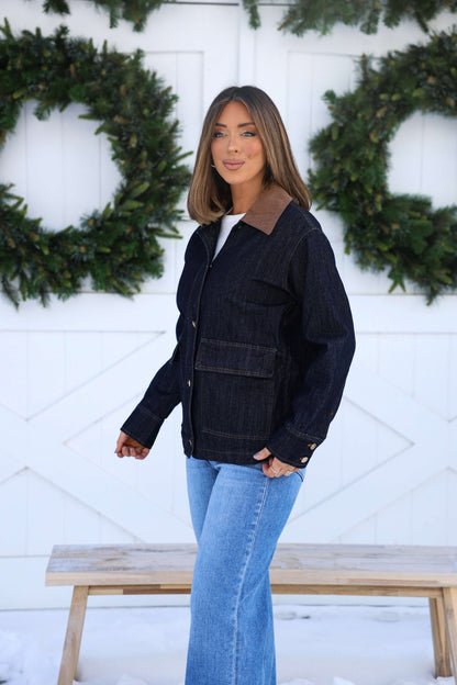 A woman in a Dark Denim Carpenter Jacket and blue jeans stands by a bench, with green wreaths on the white wall behind her.