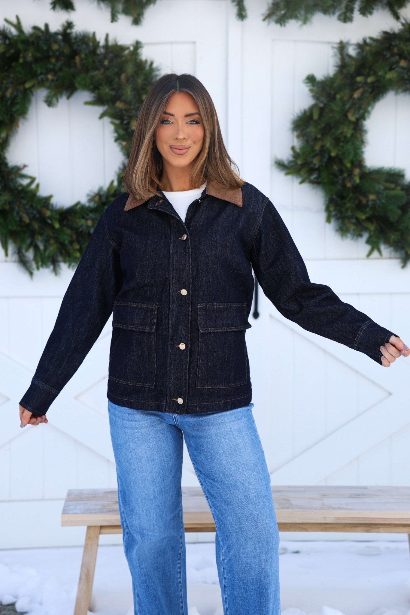 Woman smiles in front of white wall with wreaths, wearing a Dark Denim Carpenter Jacket and matching jeans.