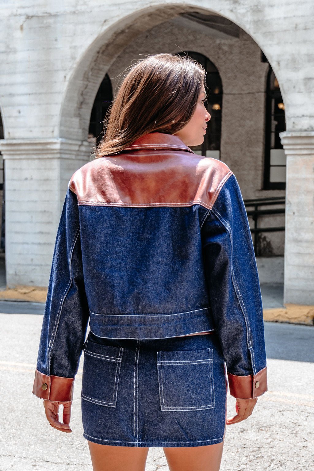 A woman stands outdoors, facing away, in the Dark Denim Contrast Button Up Jacket - DOORBUSTER with a matching denim skirt.