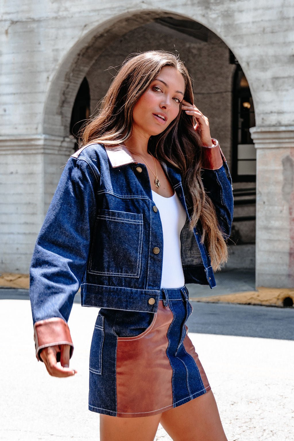 A woman wears the Dark Denim Contrast Button Up Jacket - DOORBUSTER and a brown leather skirt, outdoors, touching her hair.