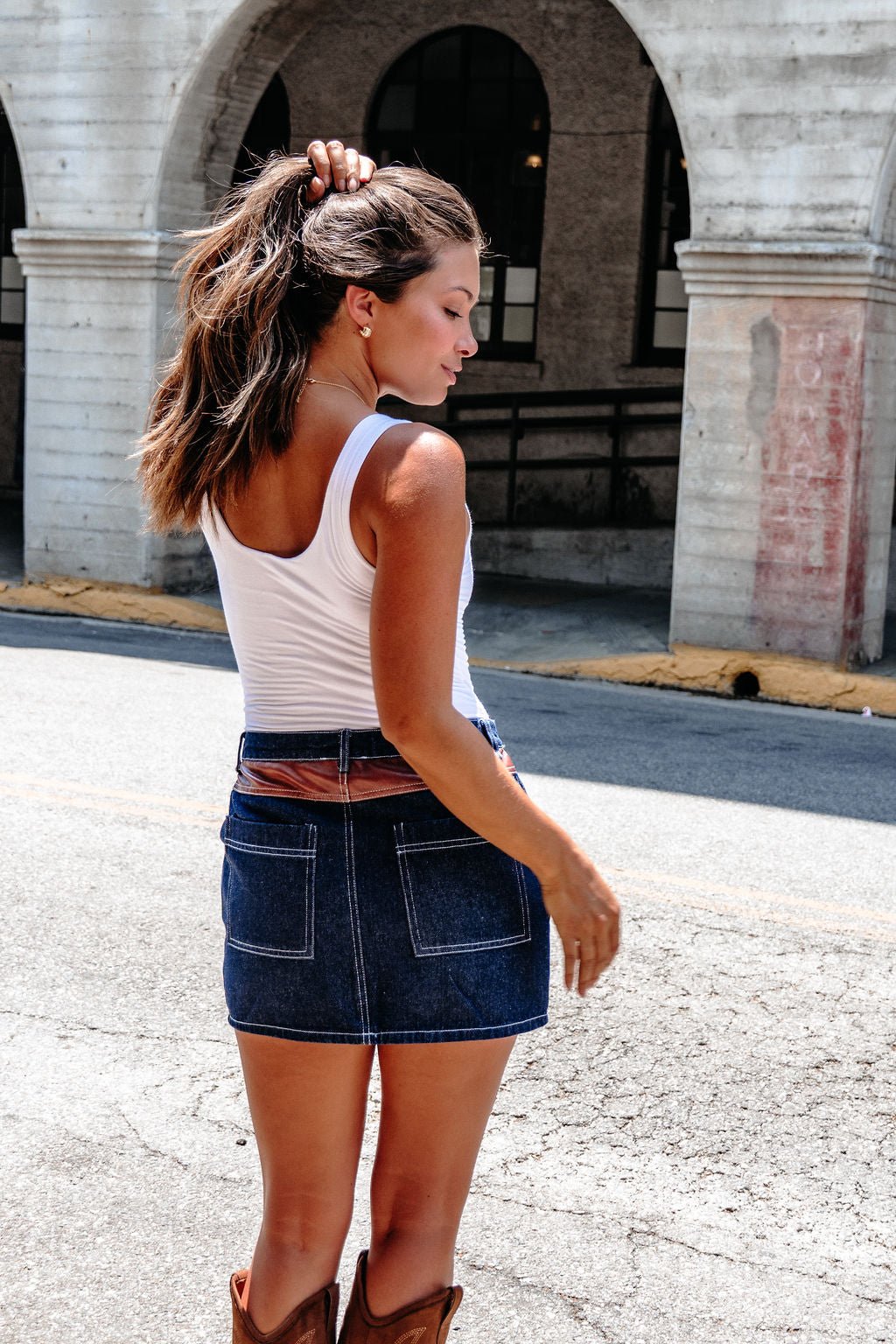 Woman in a white tank, Dark Denim Contrast Mini Skirt - FINAL SALE, and boots stands outdoors facing away, holding her hair.