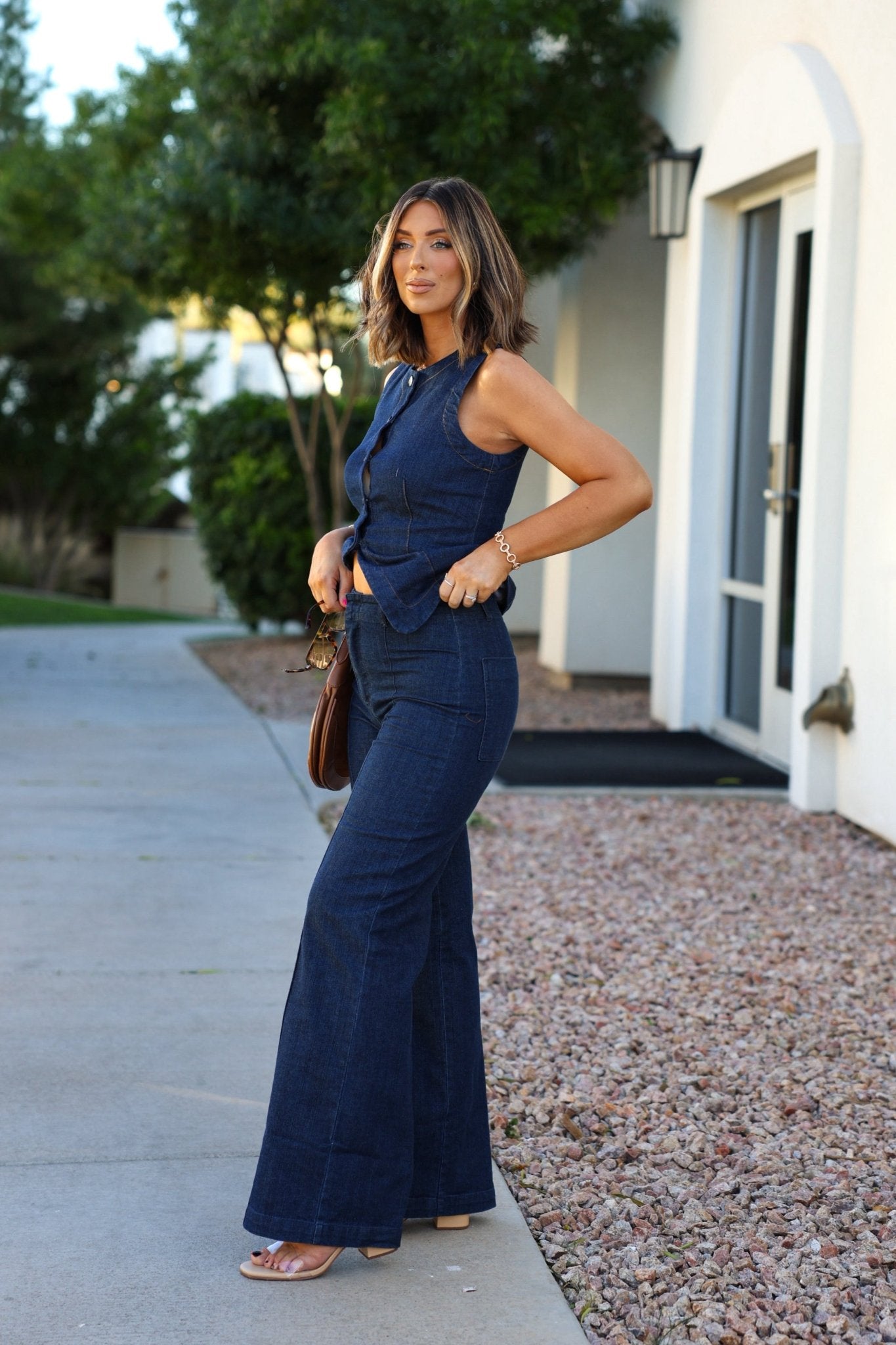 Woman outside in Dark Denim Front Seam Wide Leg Jeans, sleeveless top, holding sunglasses with trees behind.