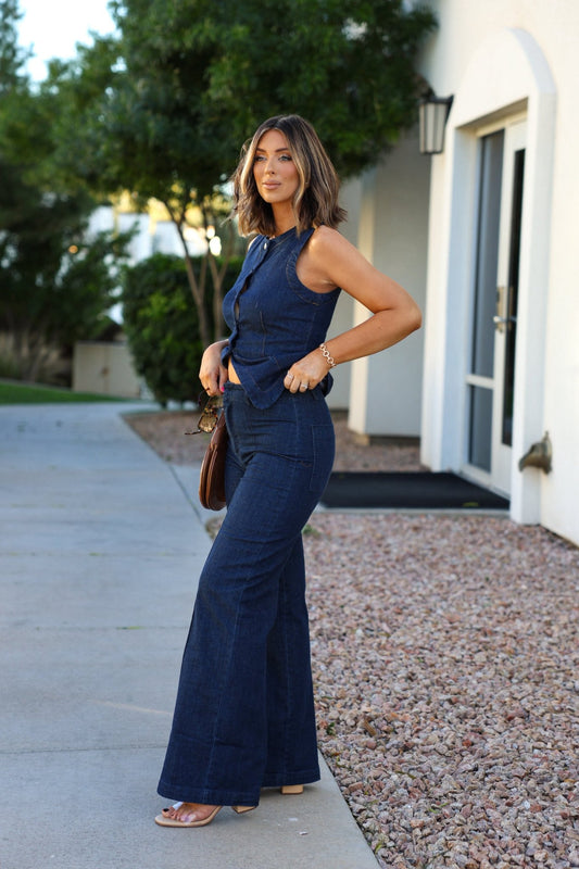 Woman outside in Dark Denim Front Seam Wide Leg Jeans, sleeveless top, holding sunglasses with trees behind.