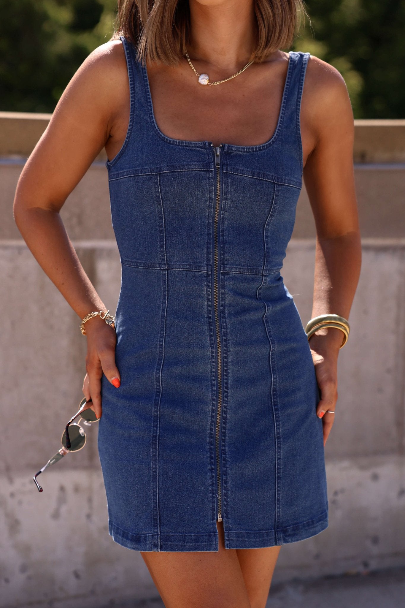 Woman outdoors in sunlight wearing the Dark Denim Zip Up Mini Dress, holding sunglasses.