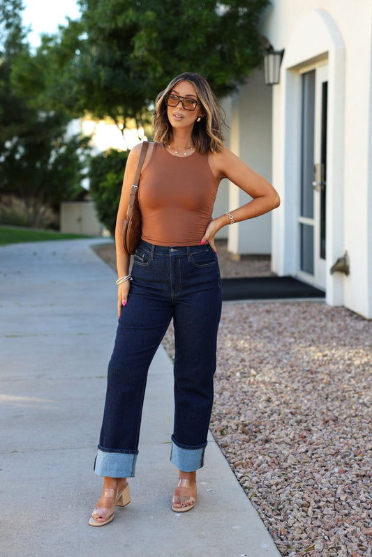 Woman wears Dark Wash High Rise Cuffed Dad Jeans, a brown sleeveless top, and heels, posing outside near a white building and trees.