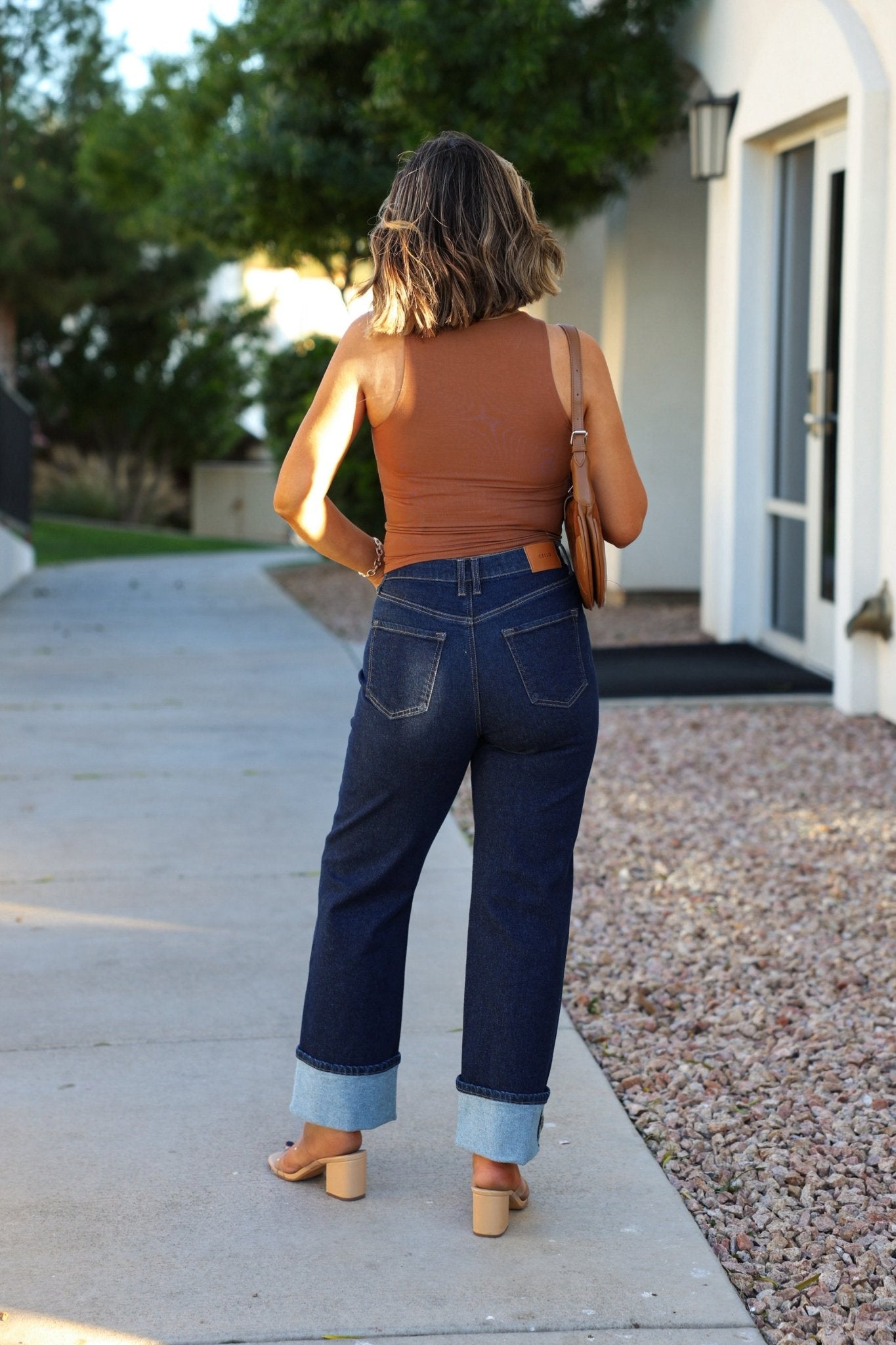 A woman with wavy hair stands outside in sandals, wearing a sleeveless brown top and Dark Wash High Rise Cuffed Dad Jeans.