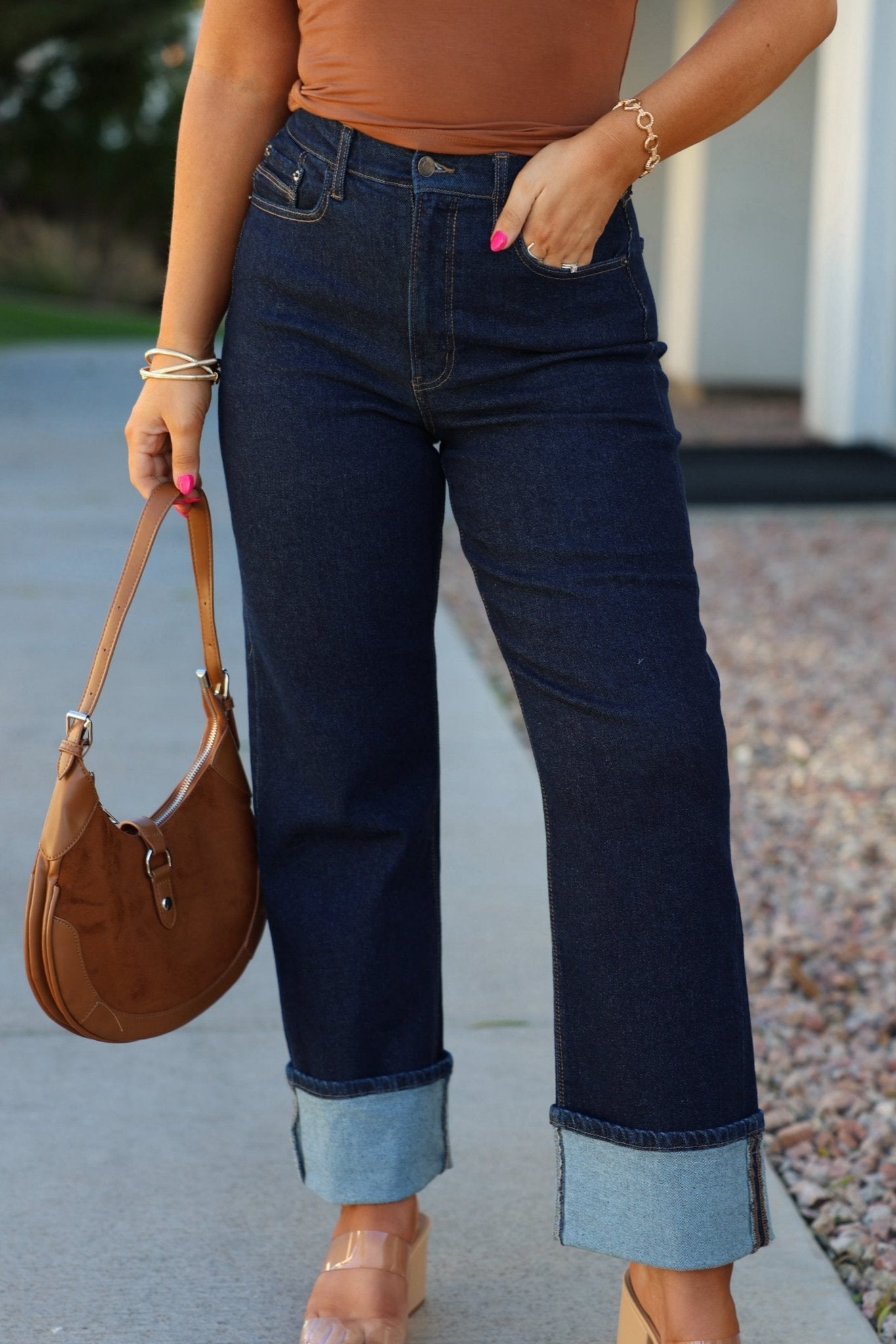 Woman in Dark Wash High Rise Cuffed Dad Jeans, tan top, brown purse, pink nails, clear heels, standing outdoors.