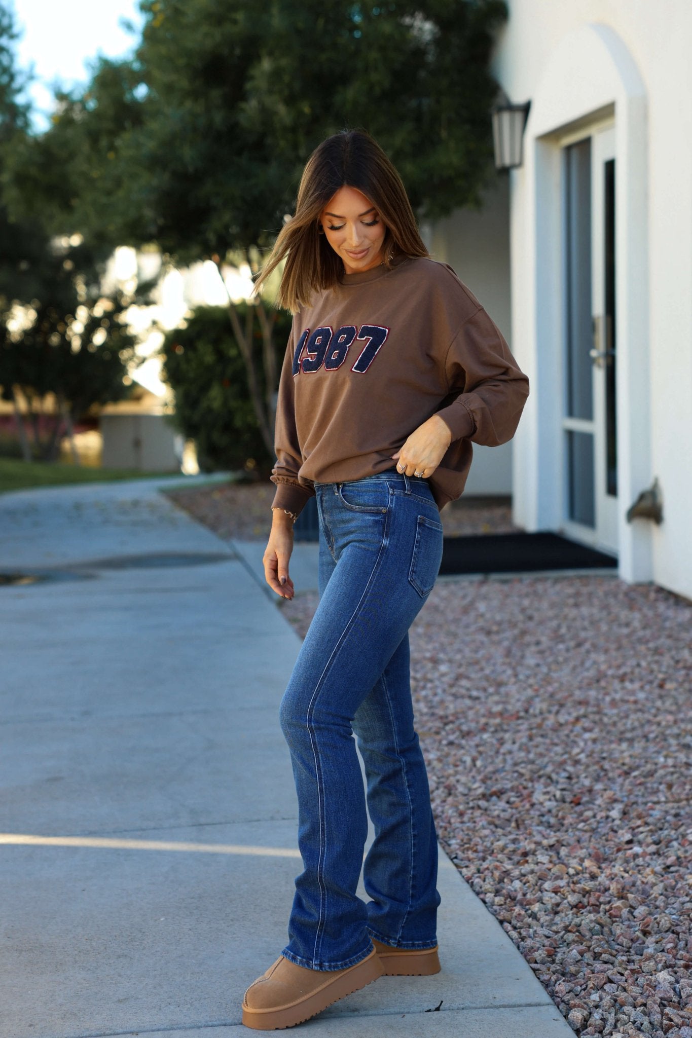 Woman outdoors in a brown 1987 sweater, tan boots, and Dark Wash Mid Rise Bootcut Jeans, looking down with a hand in her pocket.