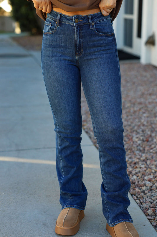Person in Dark Wash Mid Rise Bootcut Jeans and tan shoes stands on a sidewalk near a building with rocks along the side.