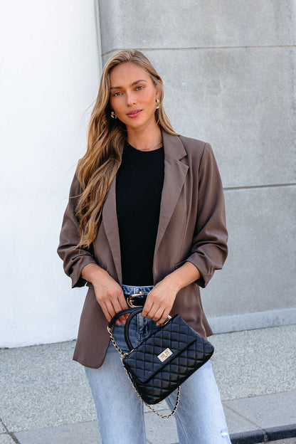 Woman in the Day To Night Blazer - Mocha with ruched sleeves, black top, jeans, and quilted purse, standing outdoors.