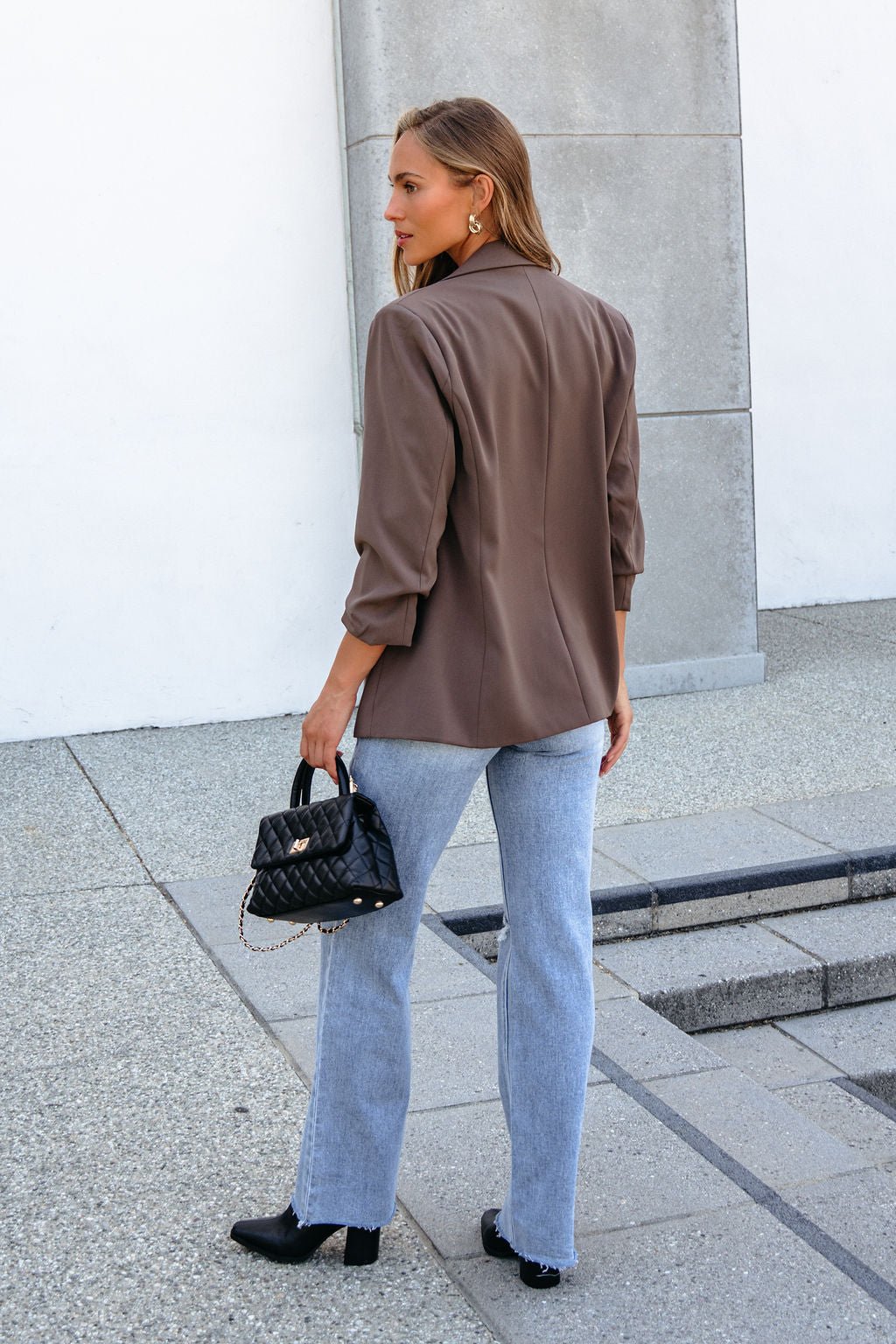 Woman in a Day To Night Blazer - Mocha, light blue jeans, and black heels stands outside with a small purse, facing away.