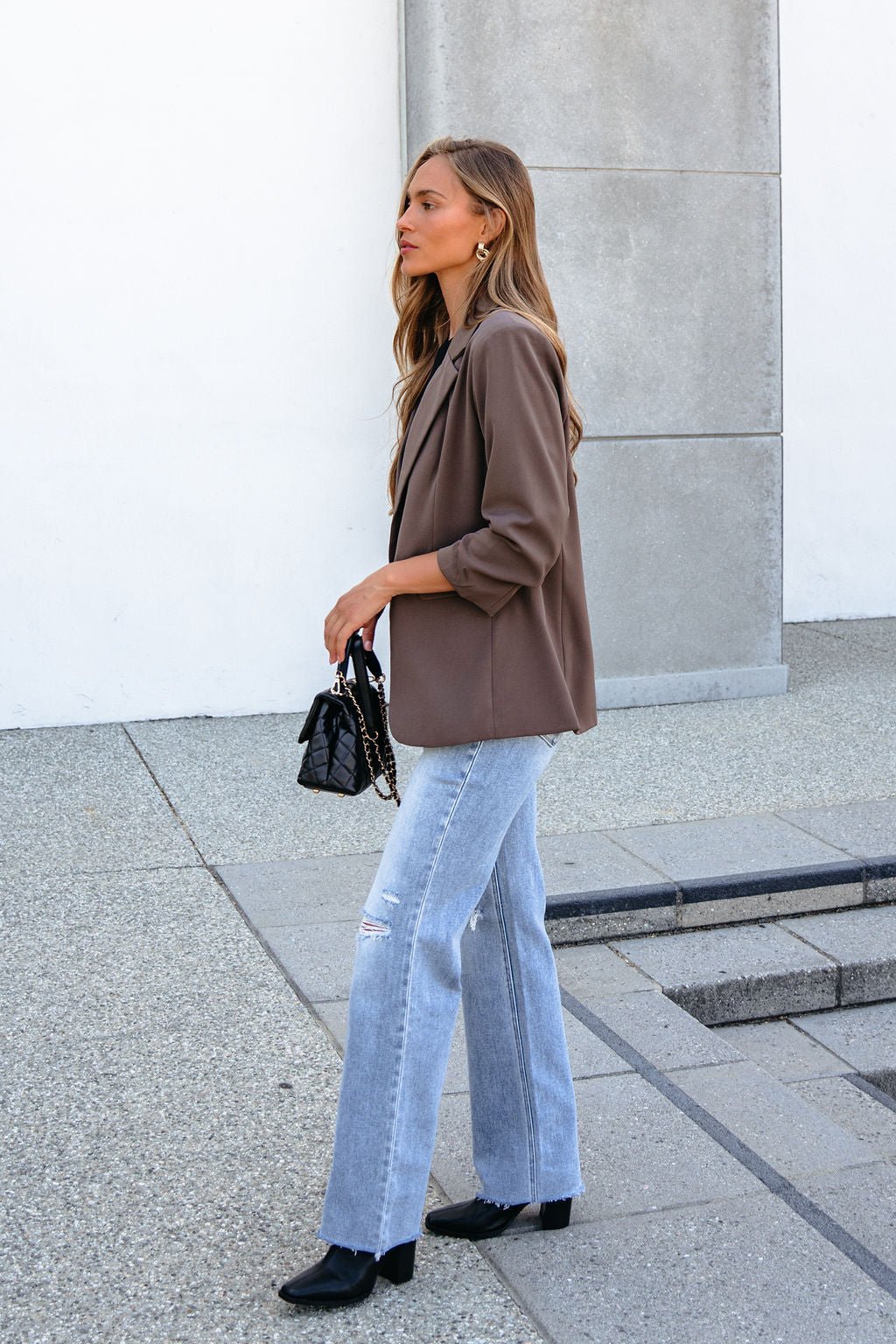 Woman in the Day To Night Blazer - Mocha, light ripped jeans, and black boots holds a black purse on a paved surface.