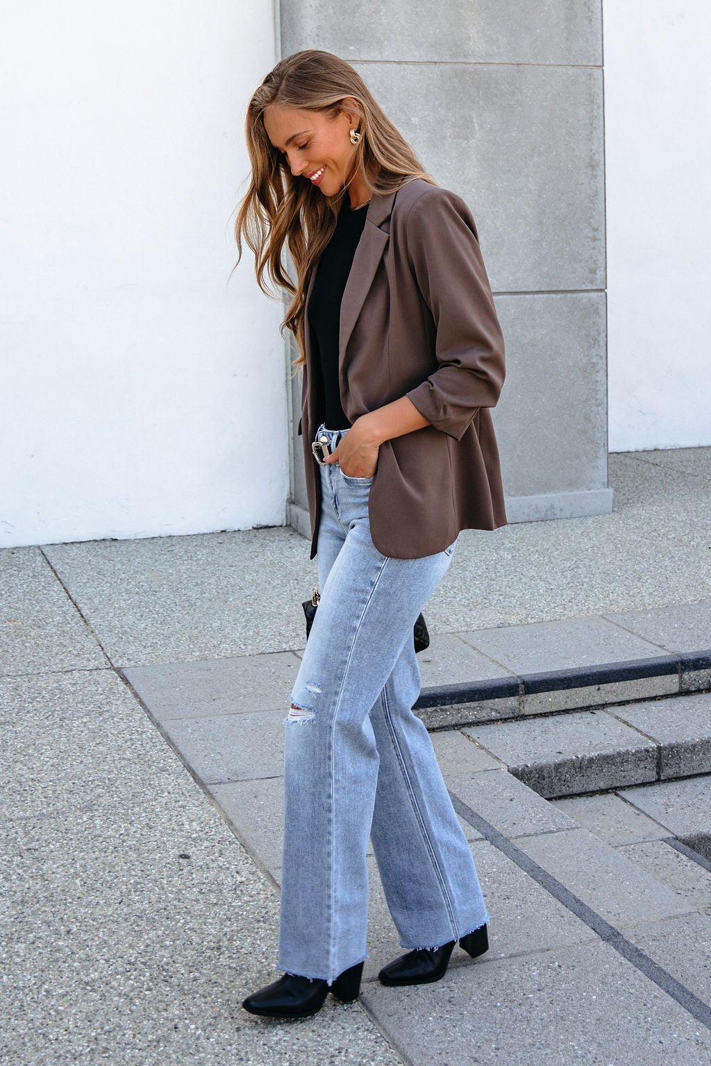 Woman in the Day To Night Blazer - Mocha, black top, and jeans walks outside smiling and looking down.