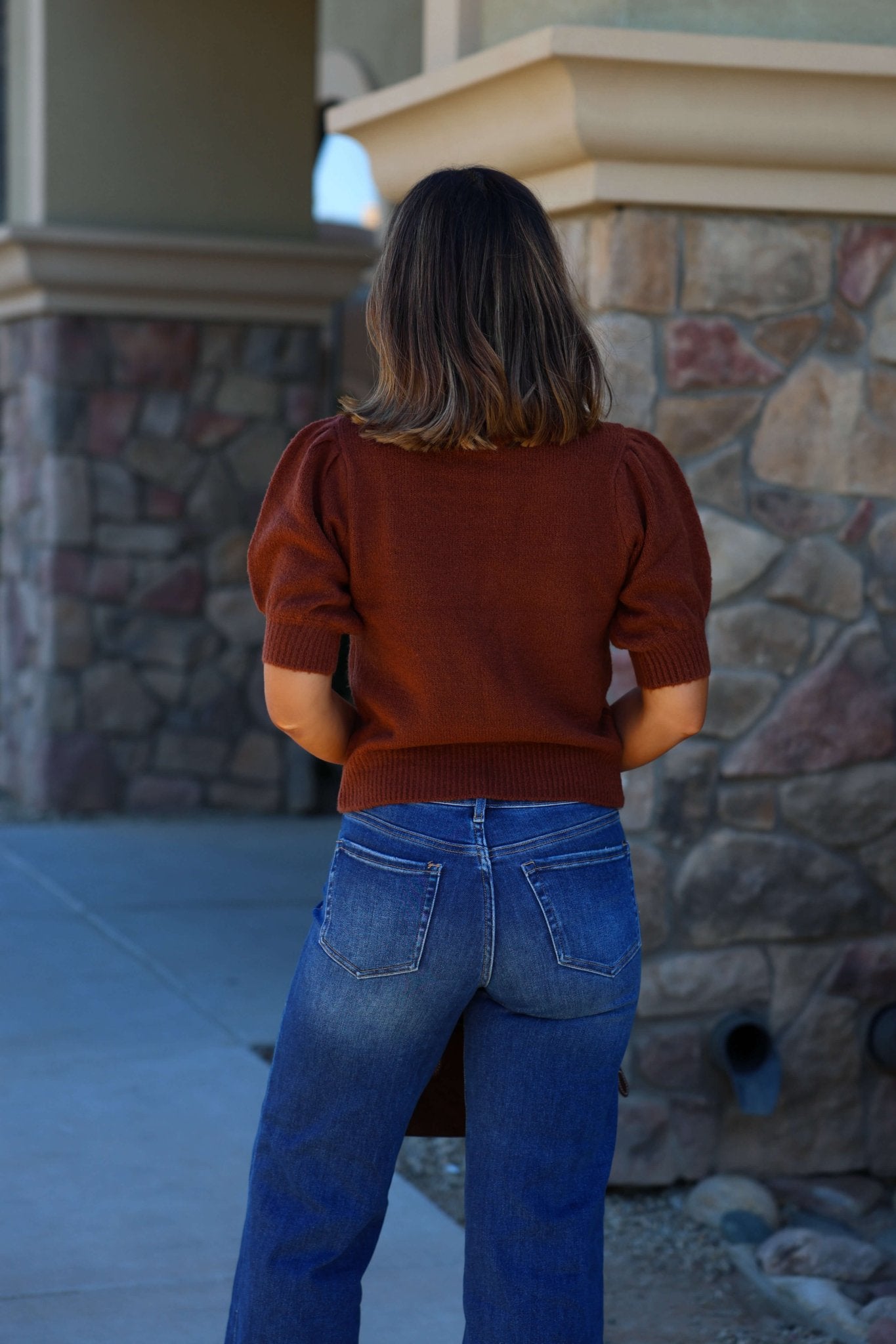A woman in the Delaney Chestnut Mock Neck Ribbed Sweater and blue jeans stands outdoors with her back to the camera near a stone wall.