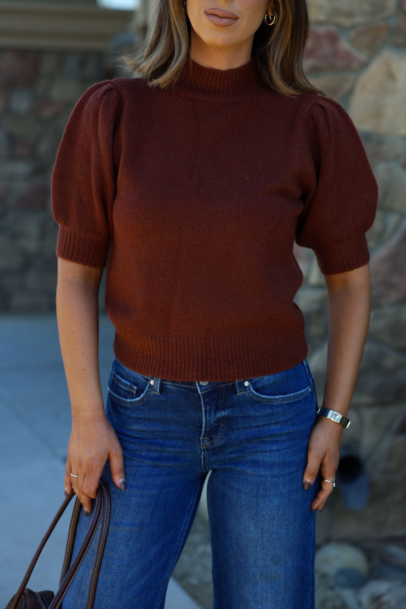 Woman in a Delaney Chestnut Mock Neck Ribbed Sweater and blue jeans holds a brown bag, outdoors by a stone wall.
