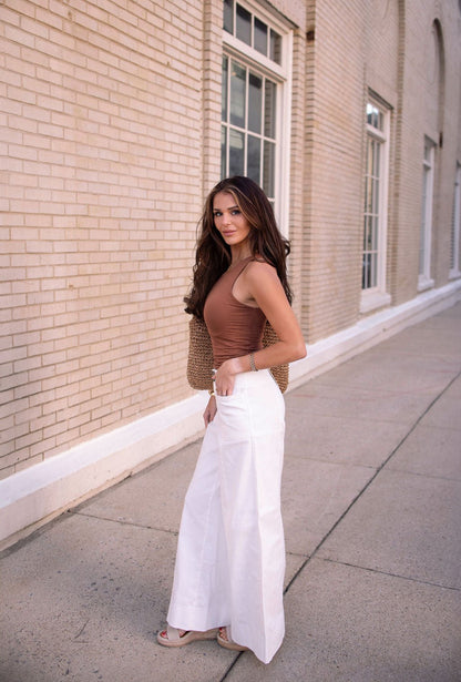 A woman in Deluxe Ivory Wide Leg Denim Pants - FINAL SALE stands on a sidewalk by a brick building, looking at the camera.