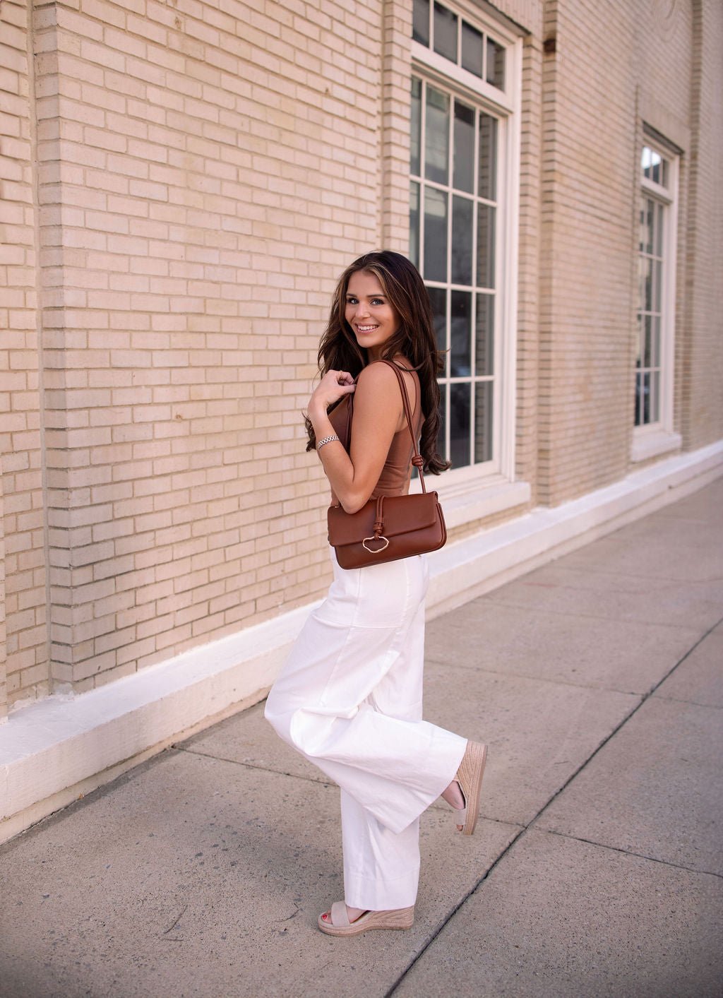 A woman smiles in Deluxe Ivory Wide Leg Denim Pants - FINAL SALE, posing by a beige brick building with large windows.