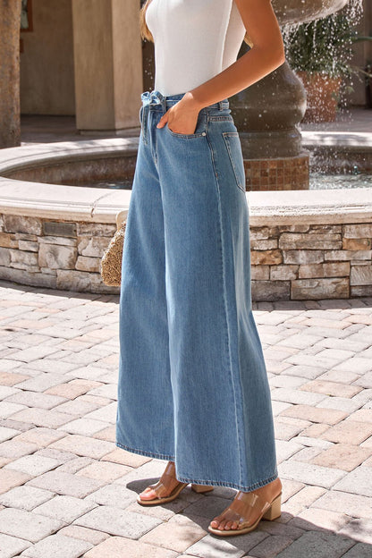 Woman wearing Denim Belted Wide Leg Pants, a white sleeveless top, and heeled sandals stands by a stone fountain.