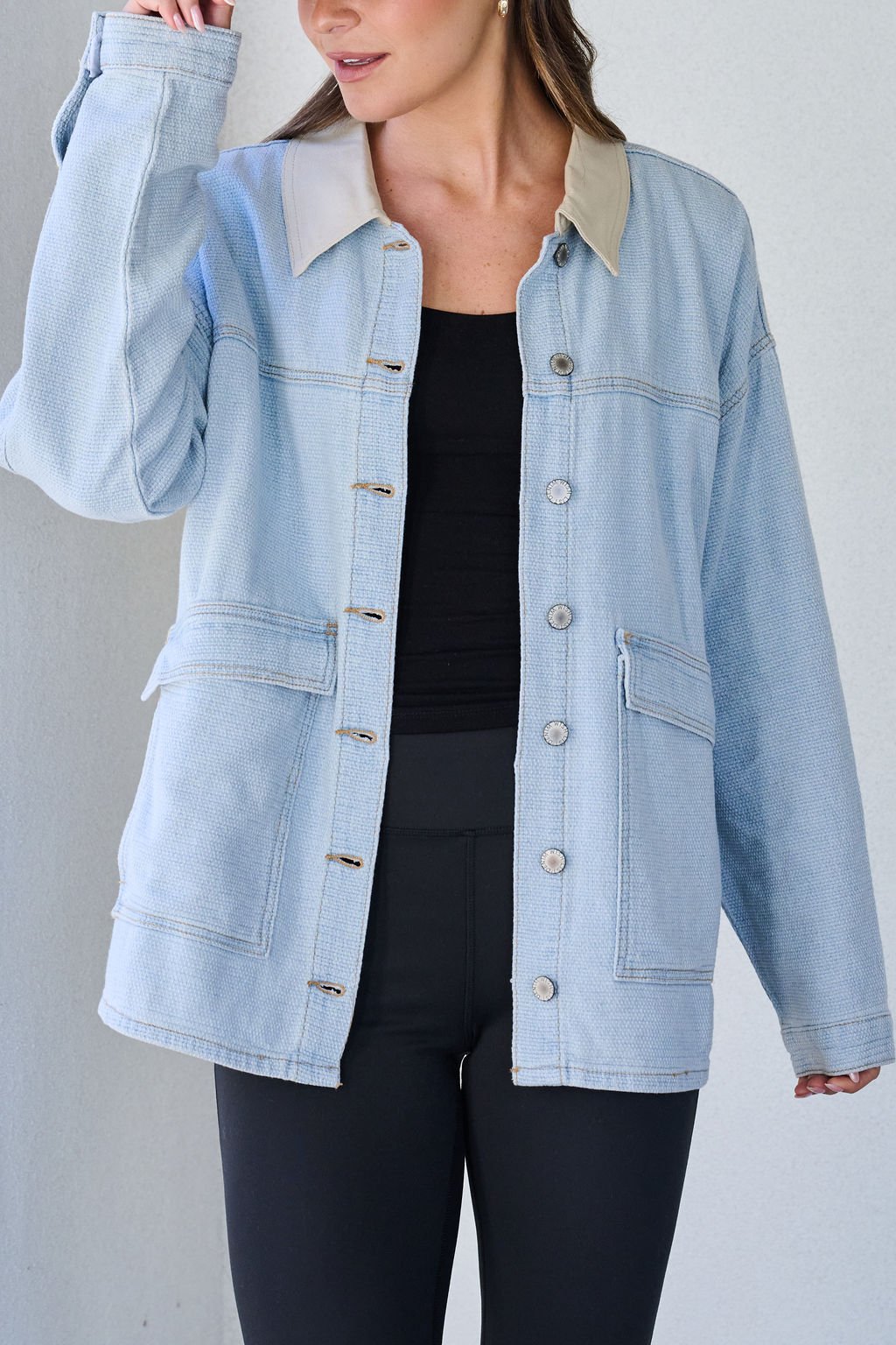 A woman wears the Denim Buttoned Utility Jacket in light blue over a black top and pants, standing against a light background.