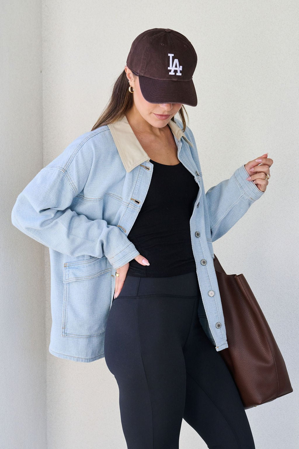 A woman in a Denim Buttoned Utility Jacket, black LA cap, black top, and leggings holds a brown tote bag while looking down.