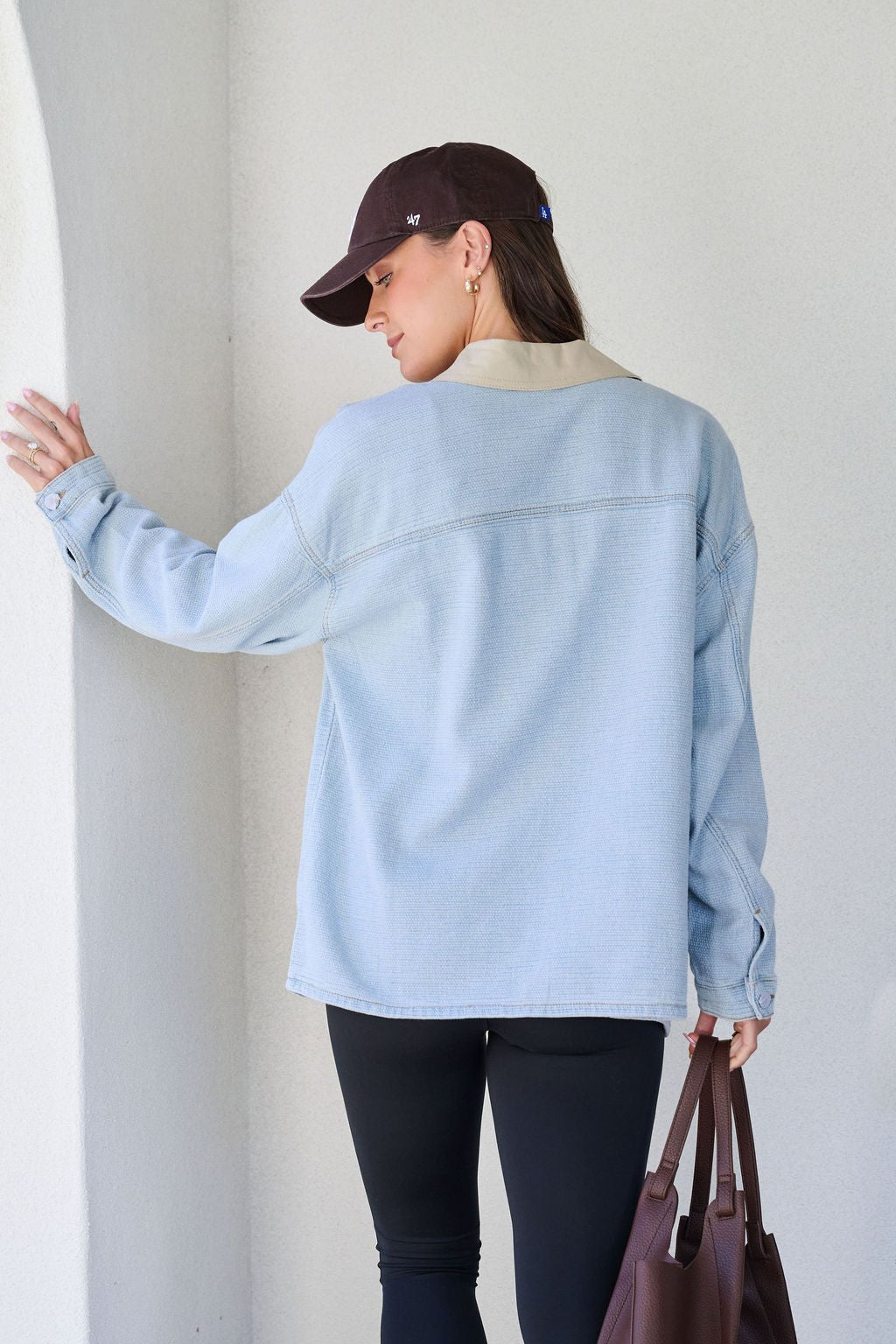Woman faces away, wearing the Denim Buttoned Utility Jacket, black cap and leggings, holding a brown tote bag.