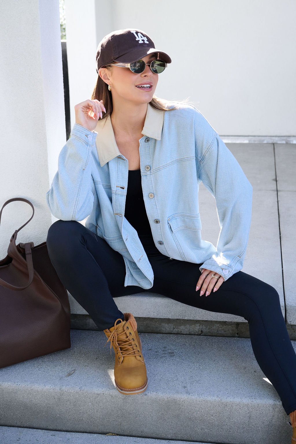 A woman wearing a Denim Buttoned Utility Jacket, black leggings, and tan boots sits on steps with a large brown bag.