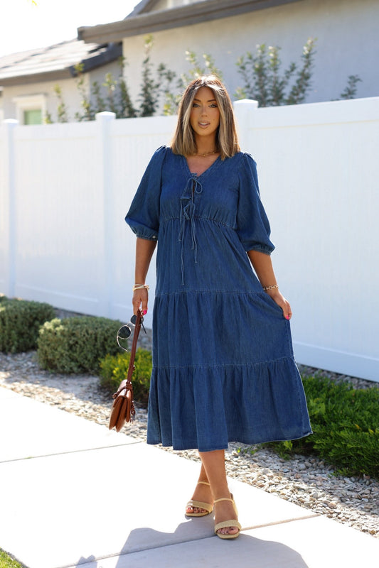 A woman in a Denim Front Tie Tiered Midi Dress stands on the sidewalk holding a brown bag and sunglasses.