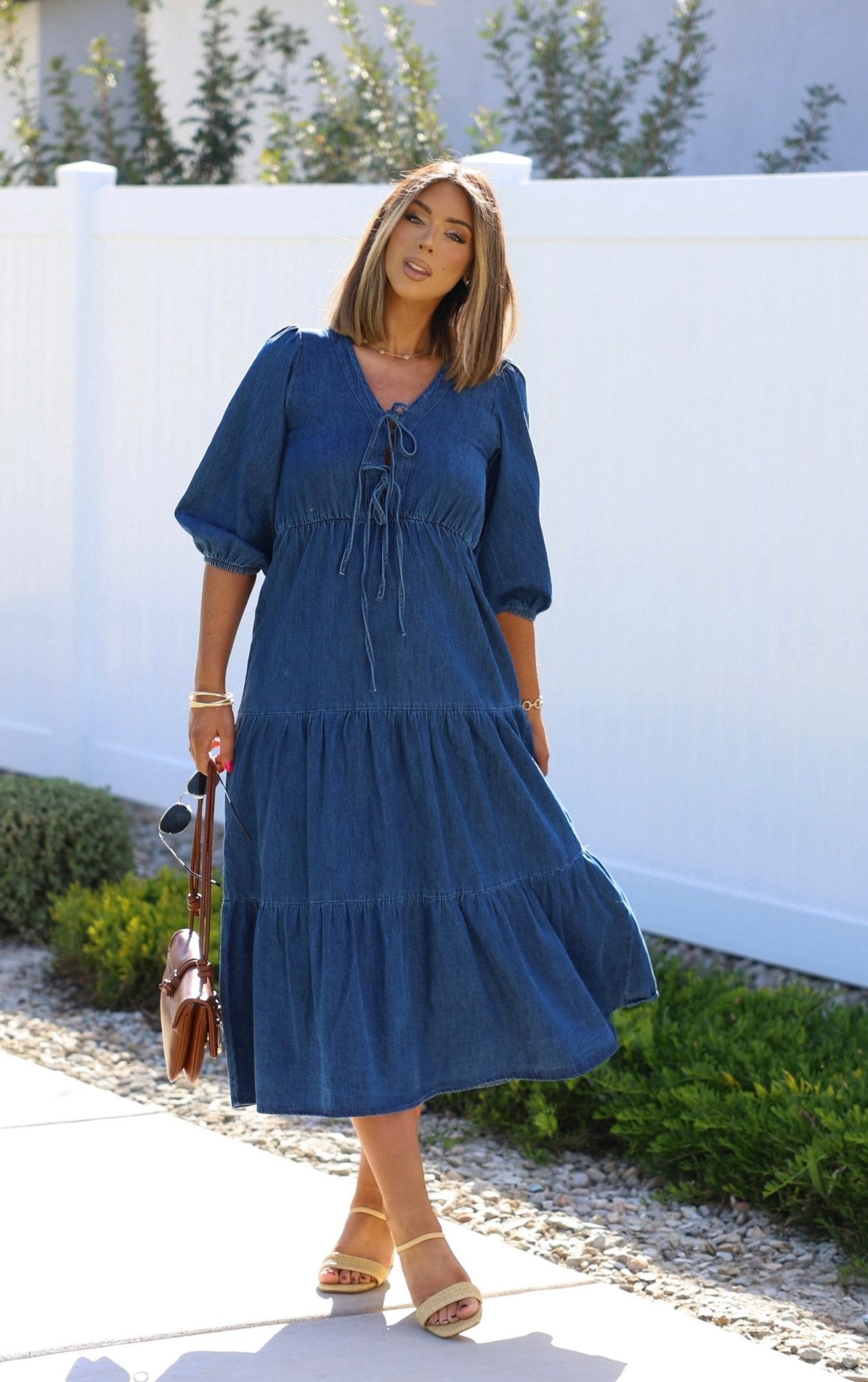 A woman in a Denim Front Tie Tiered Midi Dress stands outdoors, holding a brown handbag and wearing sandals.