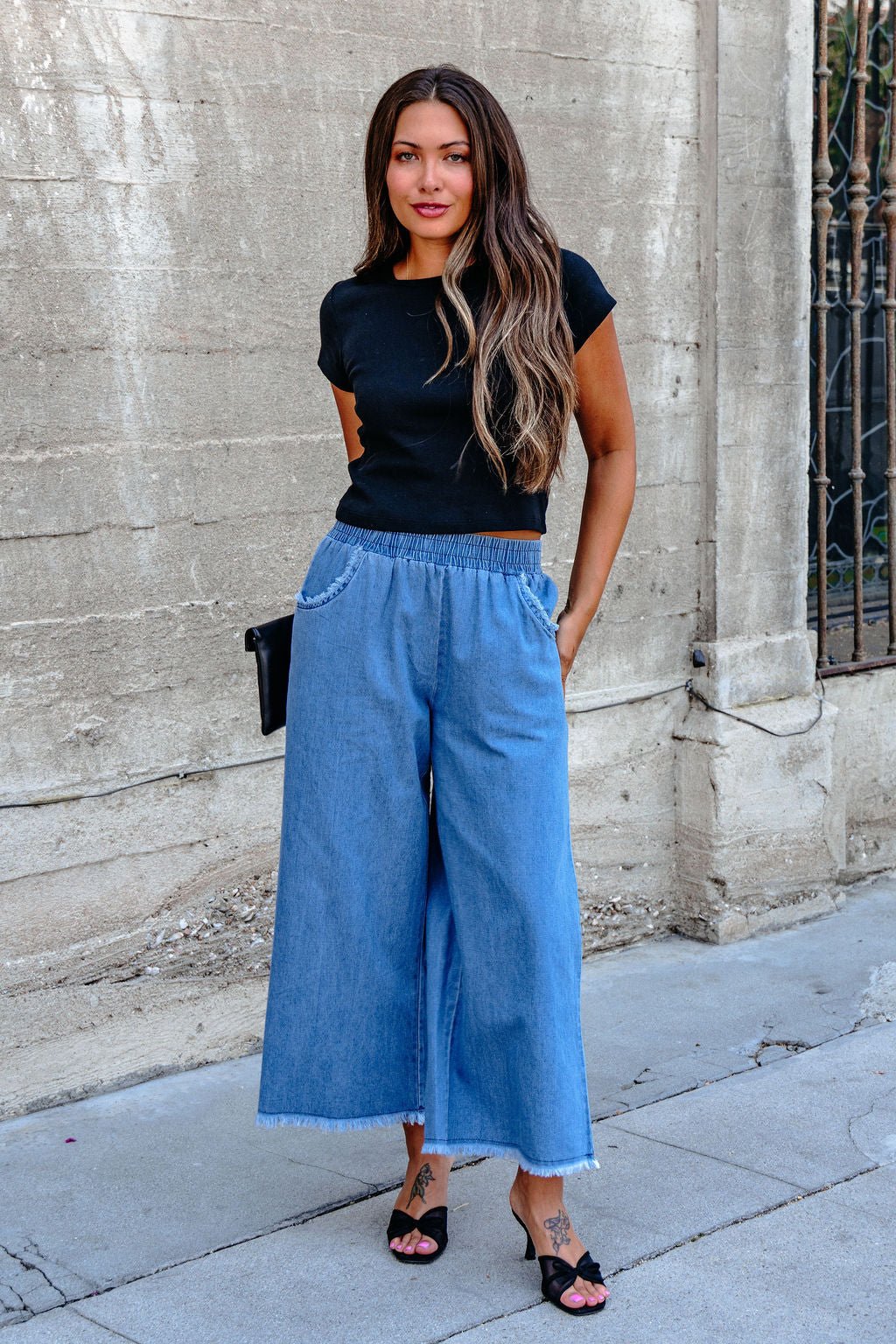 Woman outdoors in Denim Light Wash Fringe Wide Leg Pants - DOORBUSTER, black top, black heels, long hair down, hands in pockets.