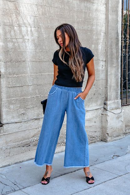 Woman in Denim Light Wash Fringe Wide Leg Pants - DOORBUSTER stands on a sidewalk by a textured concrete wall, looking down.