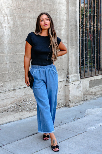 Woman with long hair wears Denim Light Wash Fringe Wide Leg Pants - DOORBUSTER, a black tee, and heels by a concrete wall.