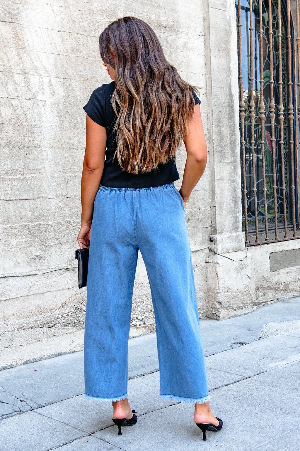 Woman with long wavy hair wears a black top and Denim Light Wash Fringe Wide Leg Pants - DOORBUSTER, facing away on the sidewalk.