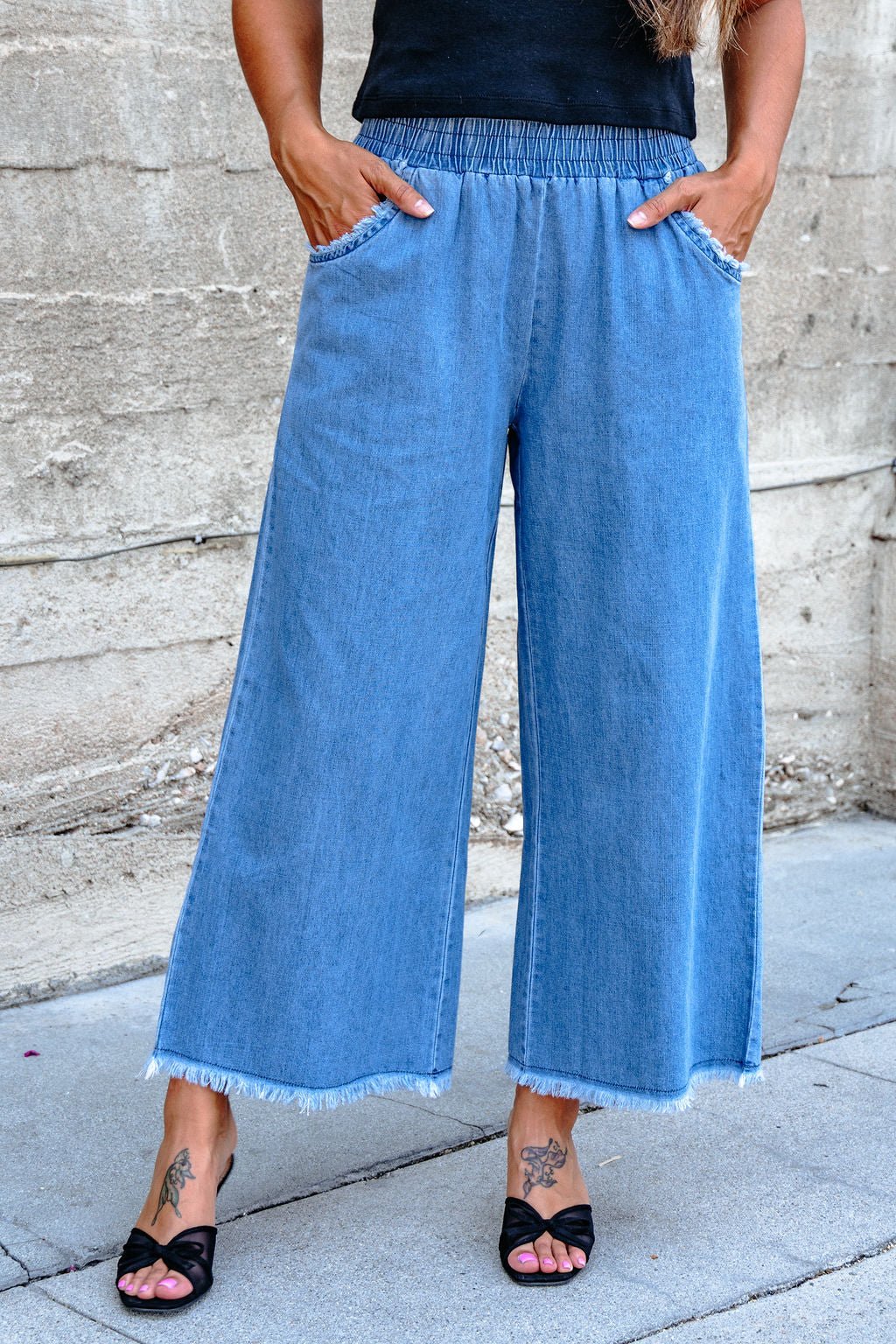 Wearing Denim Light Wash Fringe Wide Leg Pants - DOORBUSTER, black sandals, and a black top against a concrete wall.