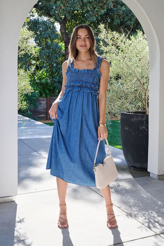 Wearing a Denim Ruffle Smocked Midi Dress and sandals, she smiles outdoors under an archway with a white handbag.