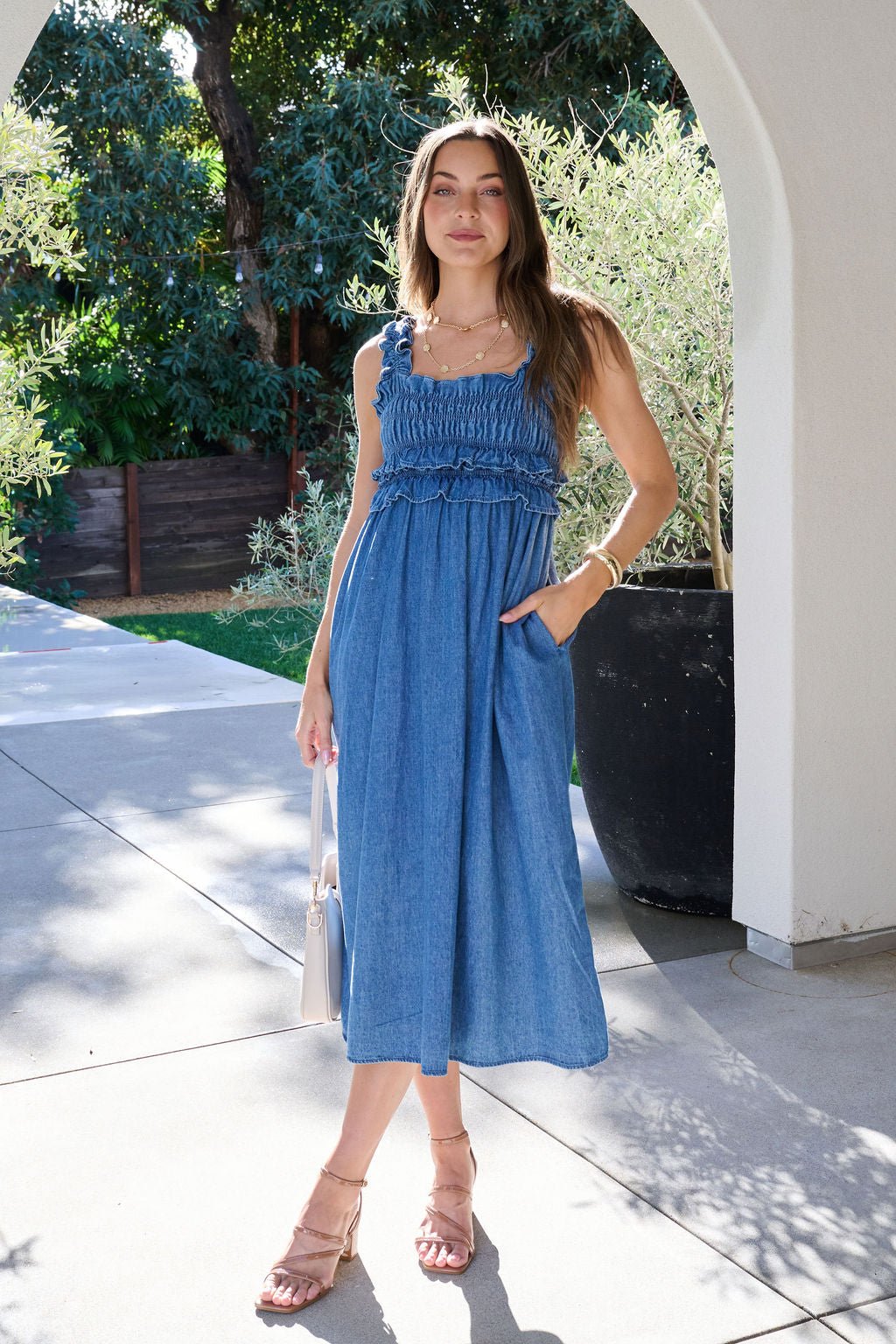 A woman wearing the Denim Ruffle Smocked Midi Dress and sandals stands outside under an archway, holding a white handbag.